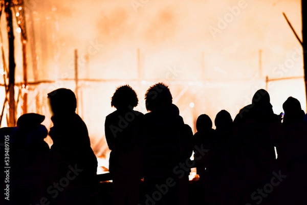 Fototapeta Dark silhouettes of people against the backdrop of a giant raging fire at night. The dark figures of a people against the backdrop of a burning building at night. People in front of the raging fire.