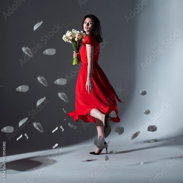 Fototapeta Full length image of young elegant woman posing in a red dress holding a bouquet of white daffodils, over grey background.