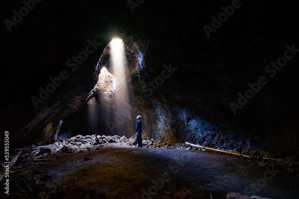 Fototapeta Adventurous athletic male standing in a lava tube looking at the sunlight shinning down into the cave through holes in the ceiling.
