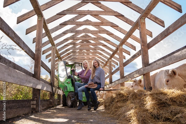 Fototapeta Portrait of two farmers women posing on farm vehicle around hay in barn.