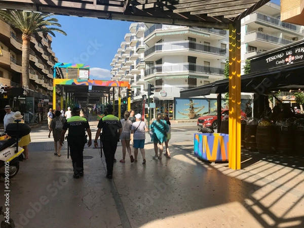 Fototapeta Salou, Spain, June 2019 - A group of people walking down a street next to a train station
