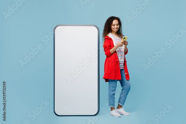 Fototapeta Full body young woman of African American ethnicity in red jacket near big huge blank screen mobile cell phone workspace copy space use smartphone isolated on plain pastel light blue cyan background.