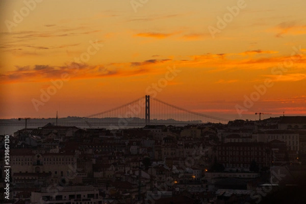 Fototapeta Lisbon city old town landscape during sunset. Golden hour in Lisbon with a view of red rooftops, blue sky and the April 25 Bridge in a distance, view of Lisbon from São Jorge Castle