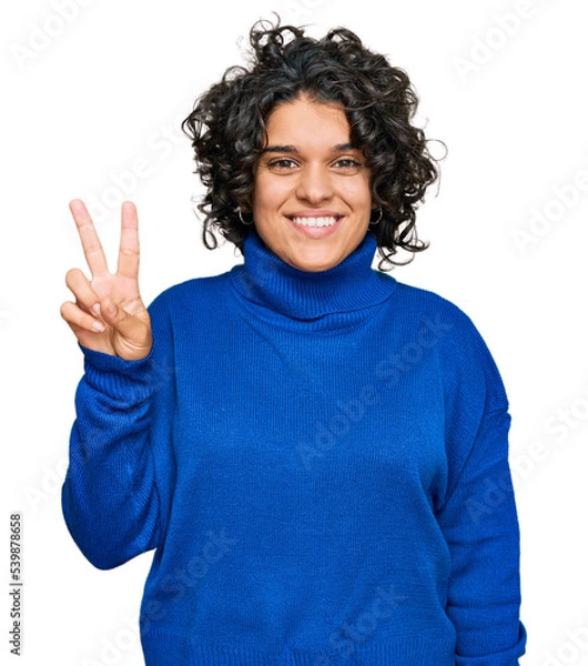 Fototapeta Young hispanic woman with curly hair wearing turtleneck sweater showing and pointing up with fingers number two while smiling confident and happy.
