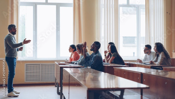 Fototapeta Friendly teacher is talking to students, smart man is raising hand and speaking sitting at table in spacious lecture hall while his classmates are writing.