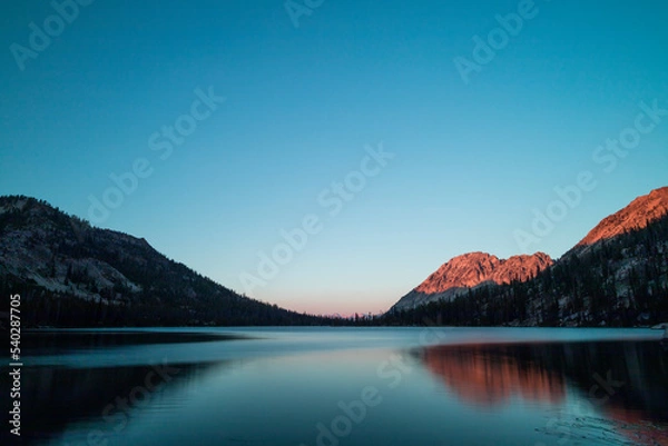 Fototapeta Toxaway Lake, located in Idaho’s Sawtooth Wilderness seen on a summer evening at sunset, with a mountain ridge illuminated reddish orange with the last rays of sunlight. The water reflects the scene.