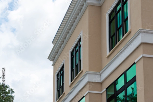 Fototapeta The exterior of a pale yellow colored wall of a house with white decorative trim and molding. The building has multiple windows with green reflective film. There's white clouds in the background. 