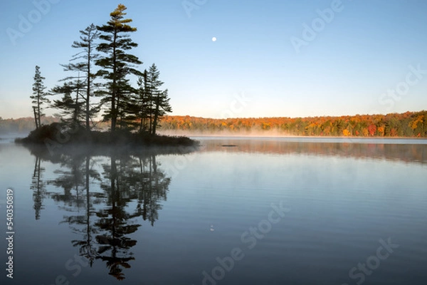 Fototapeta An island reflects on a calm lake with an autumn colored, tree lined shoreline and the moon in the background. Islet Lake, Algonquin Provincial Park, Ontario, Canada.