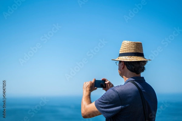 Fototapeta Caucasian man taking a picture of a landscape in San Sebastian with phone, vertical
