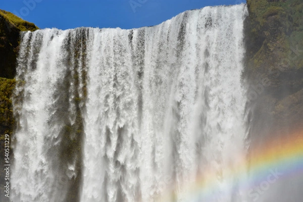 Fototapeta Splashes, water and a rainbow at the Skógafoss waterfall close up in Iceland on a summer sunny day