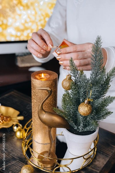 Fototapeta Unrecognizable woman featuring fire at big golden candle.Small minimalistic fir tree, festive glittering balls decorations on wooden tray. Christmas holiday celebration. New Year Xmas home hygge decor