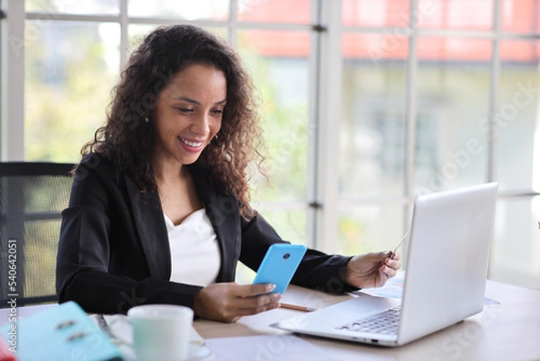 Obraz Attractive professional latin female employee worker sitting, using laptop computer with paperwork at home workplace. Businesswoman using smart mobile phone while holding credit card.