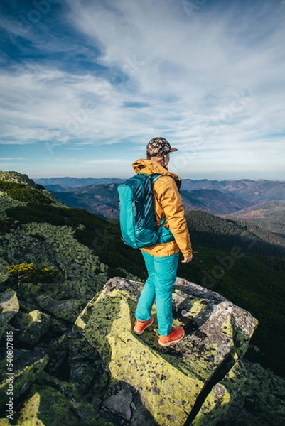Fototapeta a girl hiker in the mountains with a blue backpack walks against the background of incredibly beautiful mountains. A girl in a cap and a mustard-colored jacket walks along the rocks