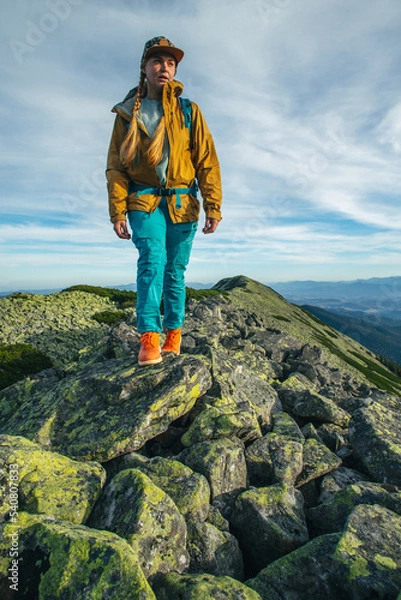 Fototapeta a girl hiker in the mountains with a blue backpack walks against the background of incredibly beautiful mountains. A girl in a cap and a mustard-colored jacket walks along the rocks