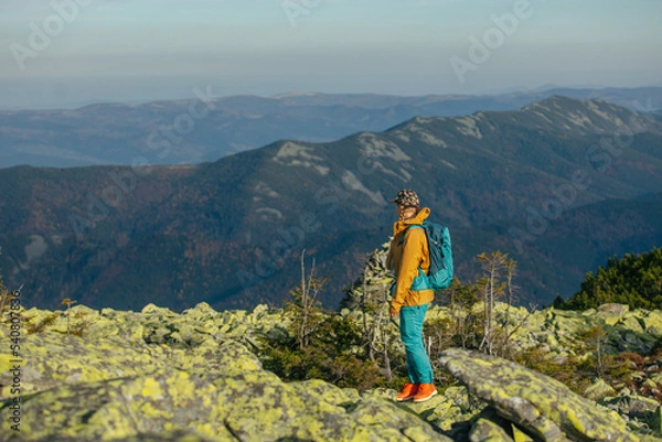 Fototapeta a girl hiker in the mountains with a blue backpack walks against the background of incredibly beautiful mountains. A girl in a cap and a mustard-colored jacket walks along the rocks