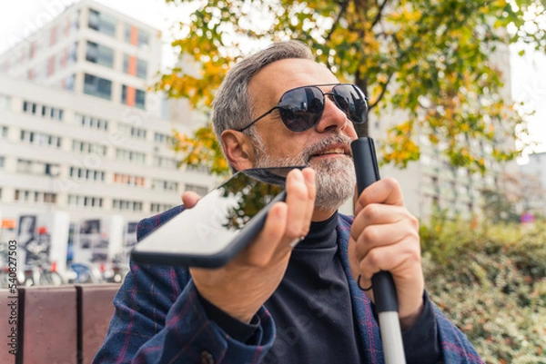 Fototapeta Blind middle-aged man with gray hair and beard wearing sunglasses holding cane and phone using text-to-speech function. Accessibilty. Modern technology. Horizontal outdoor shot. High quality photo