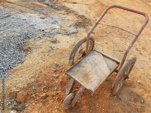 Fototapeta Wheelbarrow on construction area, Metal trolley with three wheels, Rusty old cart on the background of piles of stones and sand.