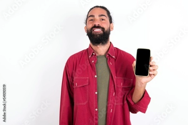 Fototapeta Smiling Caucasian man with beard wearing red shirt over white background Mock up copy space. Hold mobile phone with blank empty screen