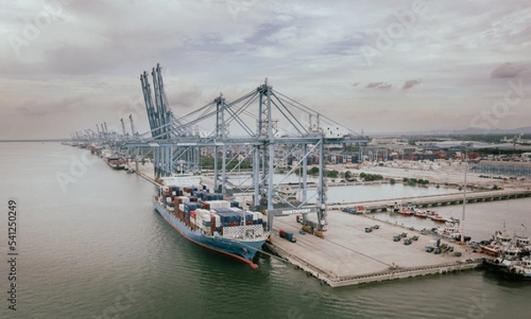 Obraz Klang, Malaysia - September 25, 2022: Cranes at the port Klang near Kuala Lumpur. Container crane at Klang Harbor. Aerial view on a container ship which is being loaded. Heavy Trucks at the Cargo bay