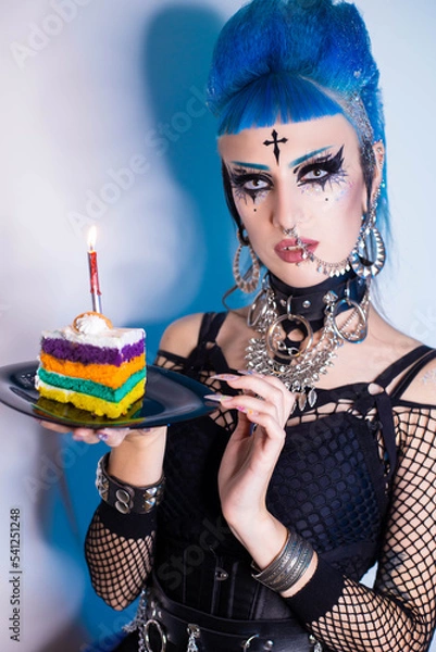 Obraz Punk girl with blue clouds, piercing and bright makeup holds a piece of colorful cake in her hands