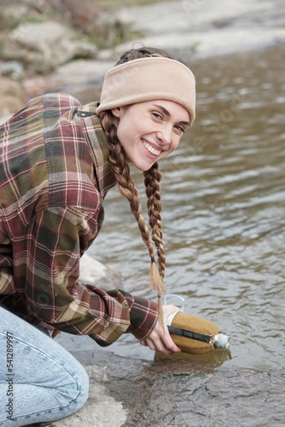 Fototapeta Woman with mountain look in the middle of autumn and filling the canteen to the river and smiling. Countryside caucasian woman with braids, shirt, plaid shirt, bandana. Excursion through the forest. 