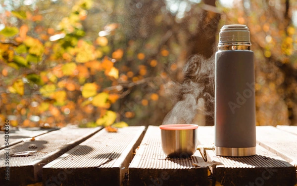 Fototapeta Thermos with mug of steaming tea on wooden table in autumn forest.