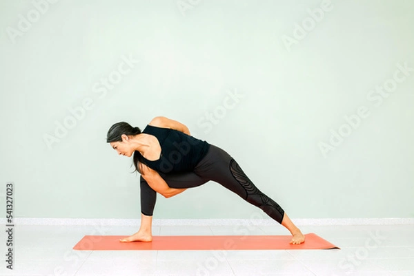 Fototapeta Hispanic mature woman performing asana exercises, workout routine. Black pants and top, studio background. Concept of yoga and wellness. Sports and healthy lifestyle.