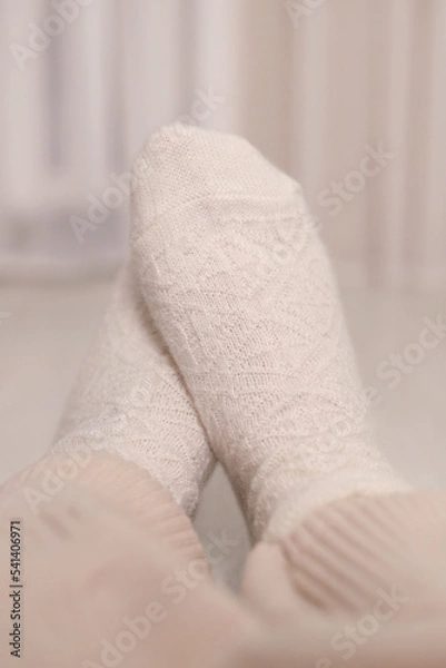 Obraz Women's feet in warm knitted woolen white socks at home under a plaid. Selective focus. Vertical photo.