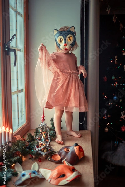Fototapeta Cute little girl is dressing up a tiny Christmas tree and trying on New Year's animal masks. Image with selective focus and toning. 