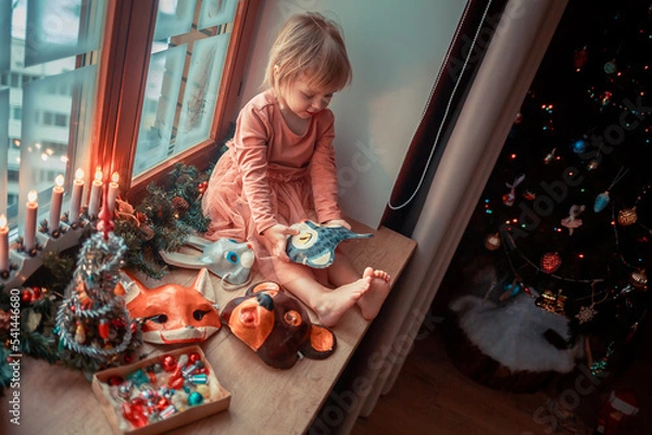 Fototapeta Cute little girl is dressing up a tiny Christmas tree and trying on New Year's animal masks. Image with selective focus and toning. 