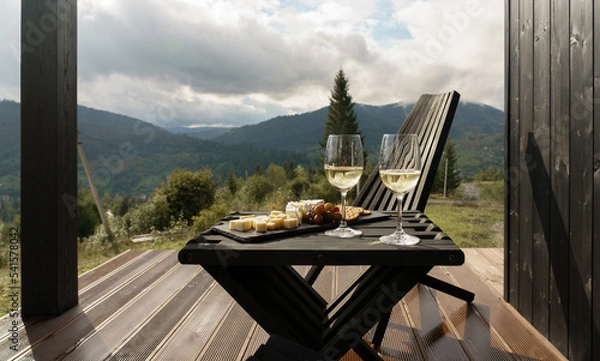 Fototapeta White wine glasses and plate of cheese on terrace with mountains view