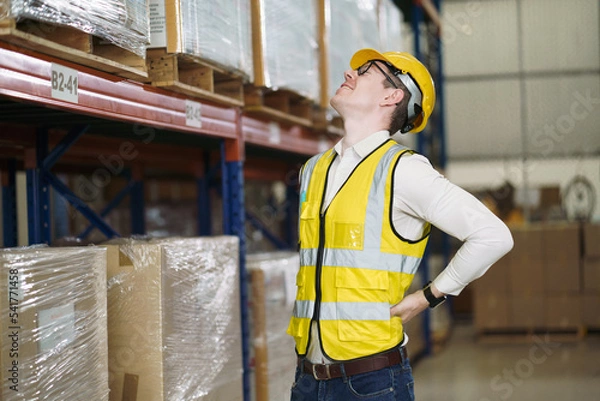Fototapeta Back ache concept of factory worker. Young man looking painful expression. Portrait of young male feeling pain while touching shoulder and his back by hand. Man suffering from office syndrome.