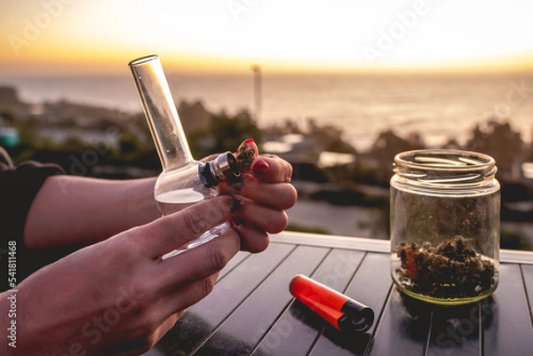 Obraz Hands with red nails putting a weed bud in a marijuana bong (weed water pipe), glass jar with marijuana buds and orange lighter with beautiful sunset sky and sea
