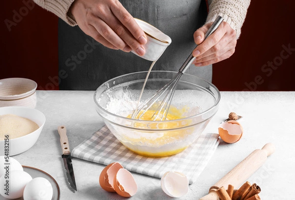 Obraz Woman pour milk into a bowl of beaten eggs and flour, mix with whisk on a gray table in the kitchen. Authentic female hands making a dough for homemade pie, Home baking concept, pie crust recipe