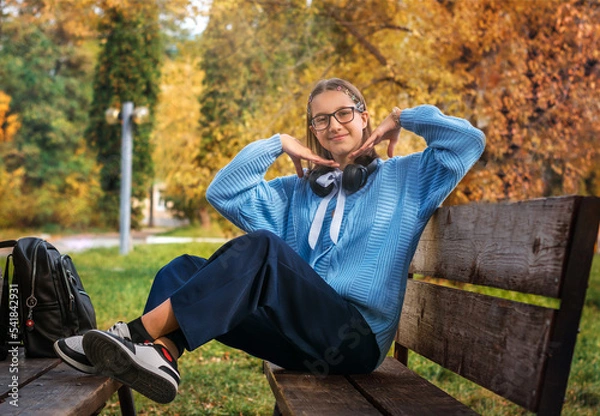 Fototapeta A young teenager with a backpack is sitting on a bench on a sunny day and smiling at the camera.