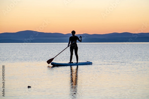 Fototapeta a young woman floats on a sapa on a mountain lake against the backdrop of sunset. Active recreation for a healthy lifestyle
