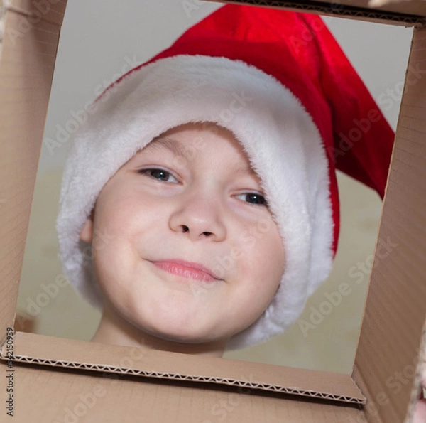 Fototapeta Little Christmas girl in a Santa hat looks into a gift box. Child is happy with a New Year's gift. The background is defocused light. The concept of Christmas and New Year 2022. High quality photo