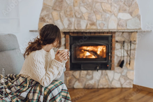 Obraz Young woman sitting at home by the fireplace with a hot tea or coffee mug and warming her hands, she is wearing white woollen sweater. Cold houses in Europe concept during energy and gas crisis.