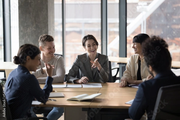 Fototapeta Happy diverse businesspeople sit at desk in office, laugh, brainstorm, discuss business ideas together. Cheerful colleagues have fun, joking enjoy communication during break at briefing in boardroom