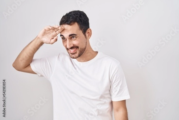 Fototapeta Handsome hispanic man standing over white background very happy and smiling looking far away with hand over head. searching concept.