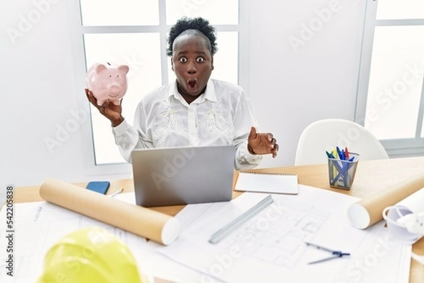 Fototapeta Young african woman working at architecture studio holding piggy bank scared and amazed with open mouth for surprise, disbelief face