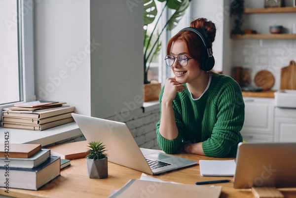Obraz Beautiful young woman in headphones looking at laptop and smiling while working from home