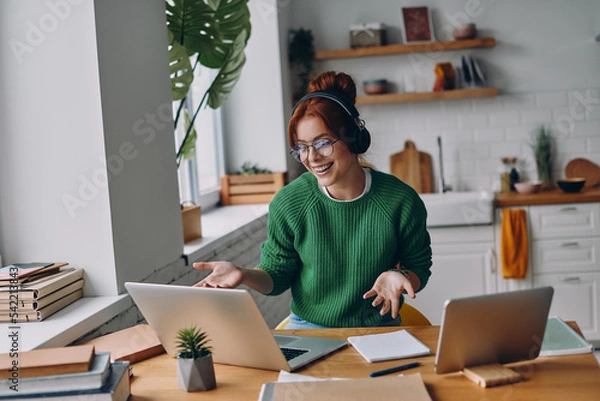 Fototapeta Cheerful young woman in headphones having web conference while working from home