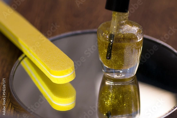 Fototapeta A bottle of translucent nail polish with gold glitter and a yellow nail file on a mirror surface