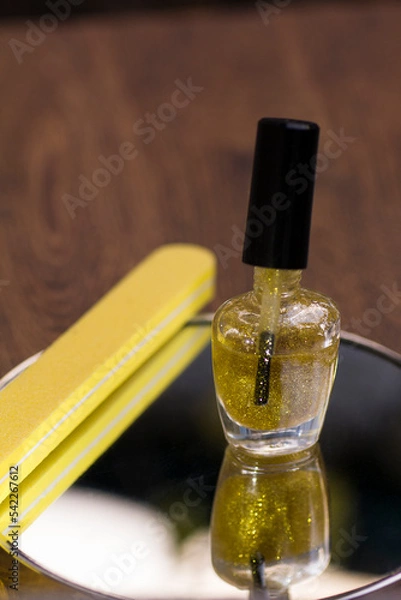 Fototapeta A bottle of translucent nail polish with gold glitter and a yellow nail file on a mirror surface