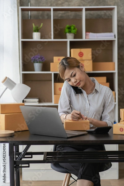 Fototapeta 
Smiling Asian woman looking at camera and using laptop to check package delivery information before sending it to customer. Small sme business entrepreneur. Online selling concept.