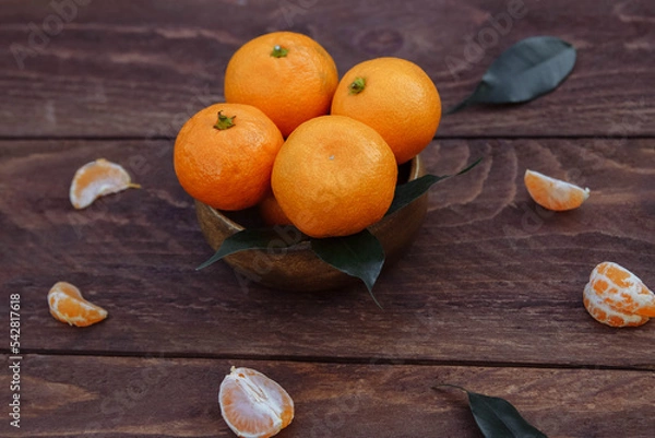 Fototapeta Ripe tangerines in a bowl on a brown wooden background close-up with green leaves in rustic style, the concept of organic healthy food, harvest season, vitamin C ingredient for dessert