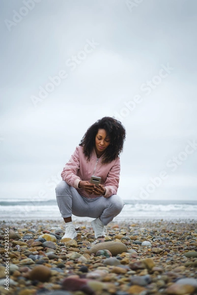 Obraz Młoda modna sportowa kobieta z fryzur afro wysyła SMS -y na smartfonie na plaży. Moda w sezonie jesiennym.