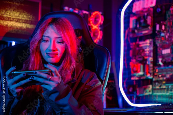 Fototapeta Young woman live stream she play video game at home neon lights living room, Gamer playing online game application on mobile smart phone wear gaming headphones, E-Sport concept