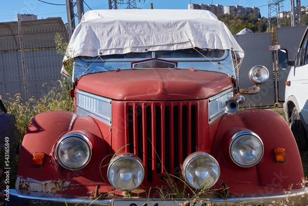 Fototapeta An old red car is parked in the grass. The car is covered with a white cloth from the sun. You can see chrome parts, headlight, bumper, hood, horn. Selective focus.